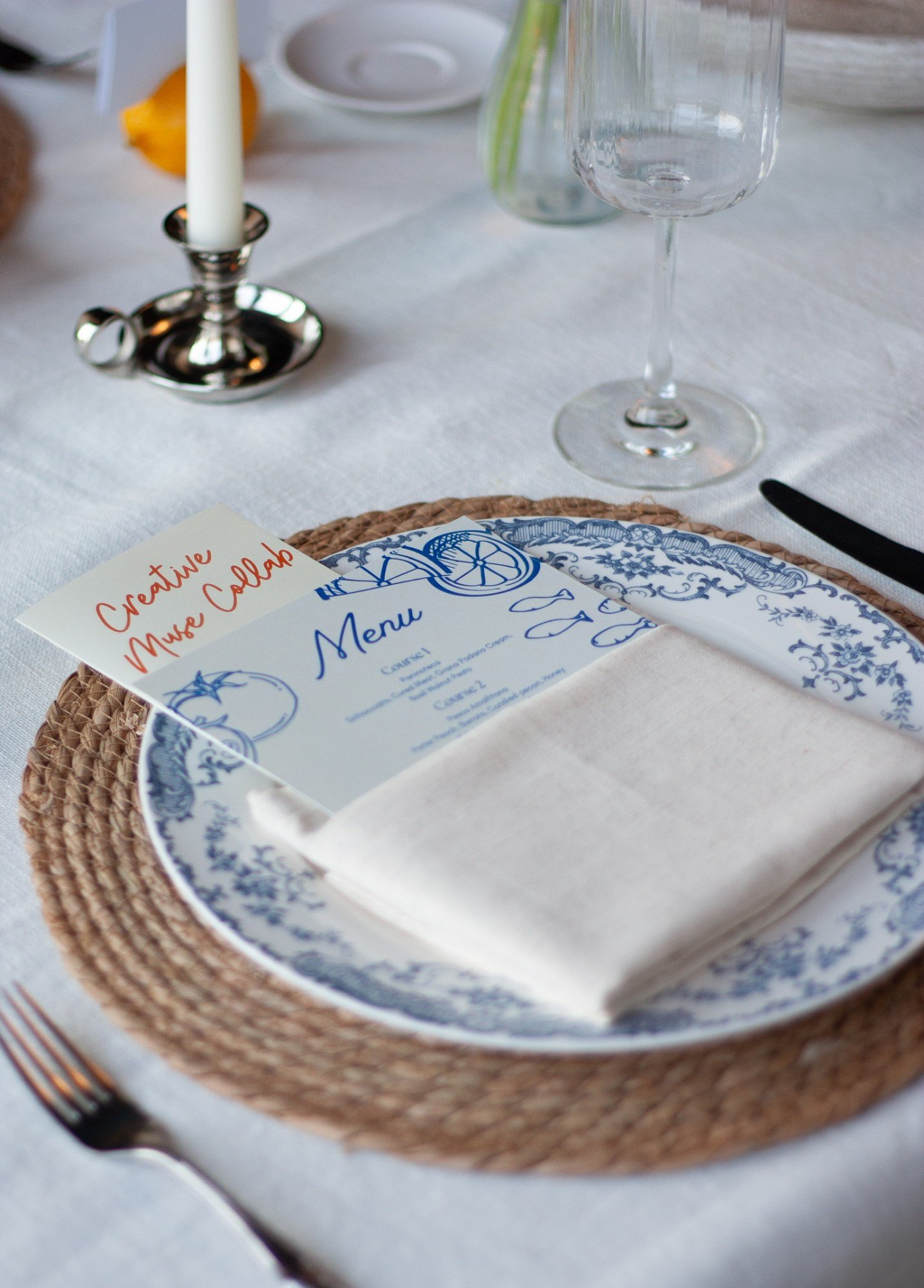 The illustrated menu and Creative Muse Collab card tucked into a linen napkin on blue and white china