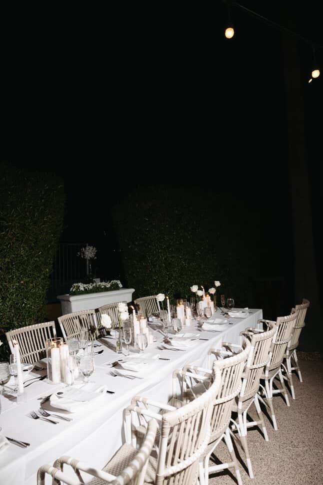 The long white table glowing with pillar candles and roses against the night hedge