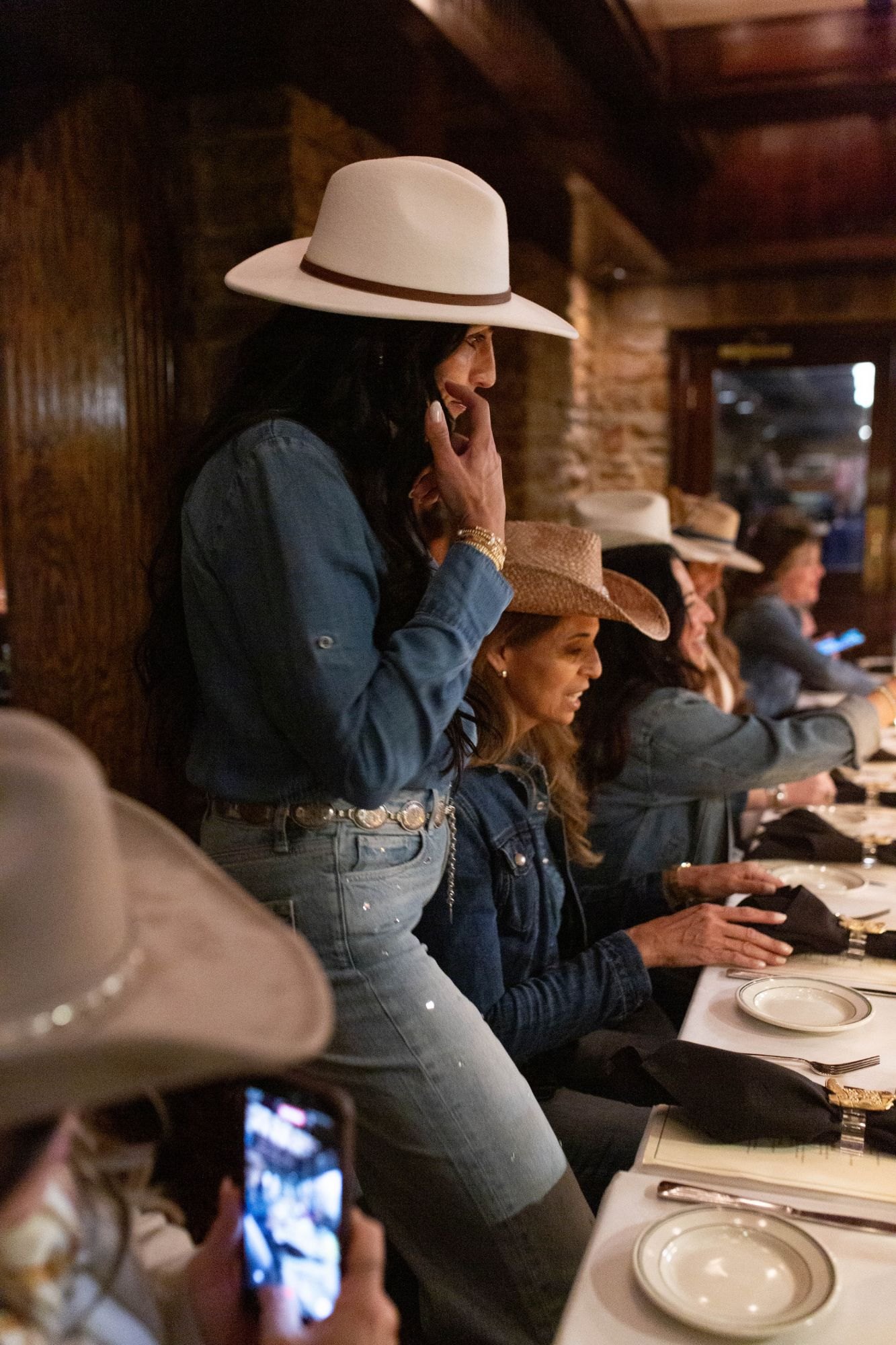 A guest in a white hat and denim stands to speak at the table