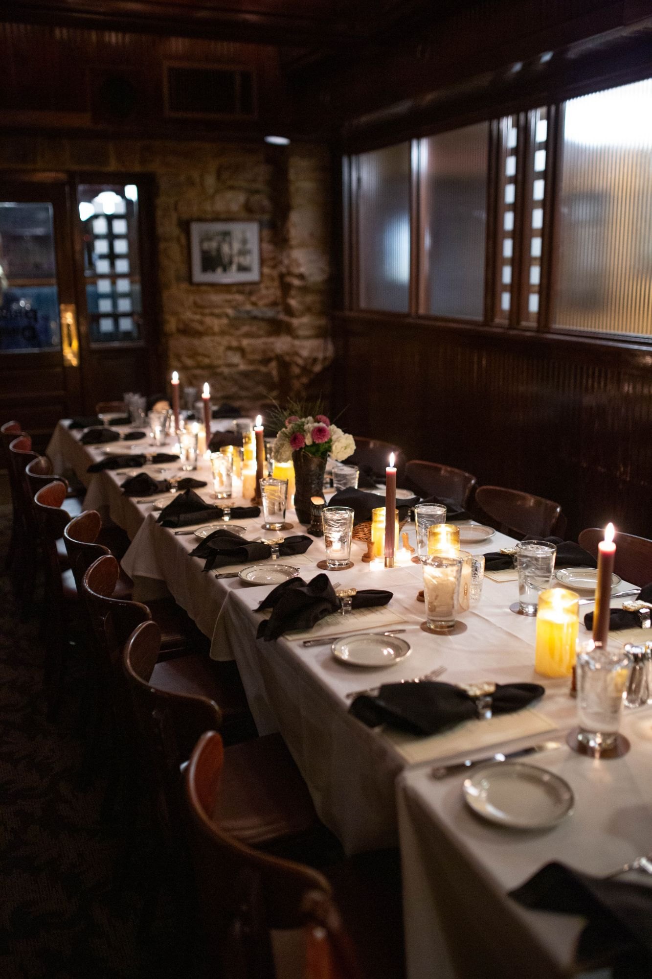 A long candlelit table with black napkins and florals in a wood-paneled private room