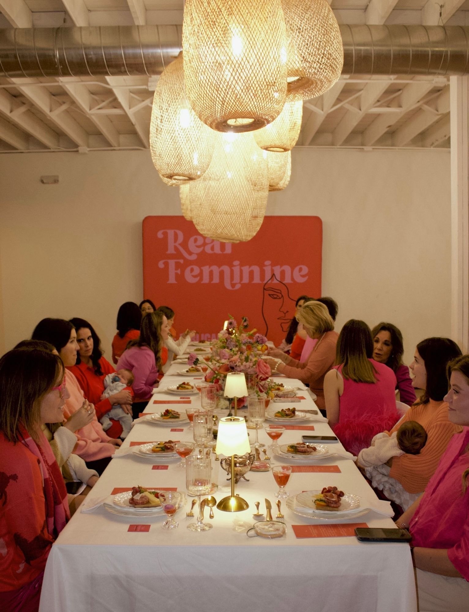 Guests in pinks and reds dine beneath rattan pendants in front of the Real Feminine backdrop
