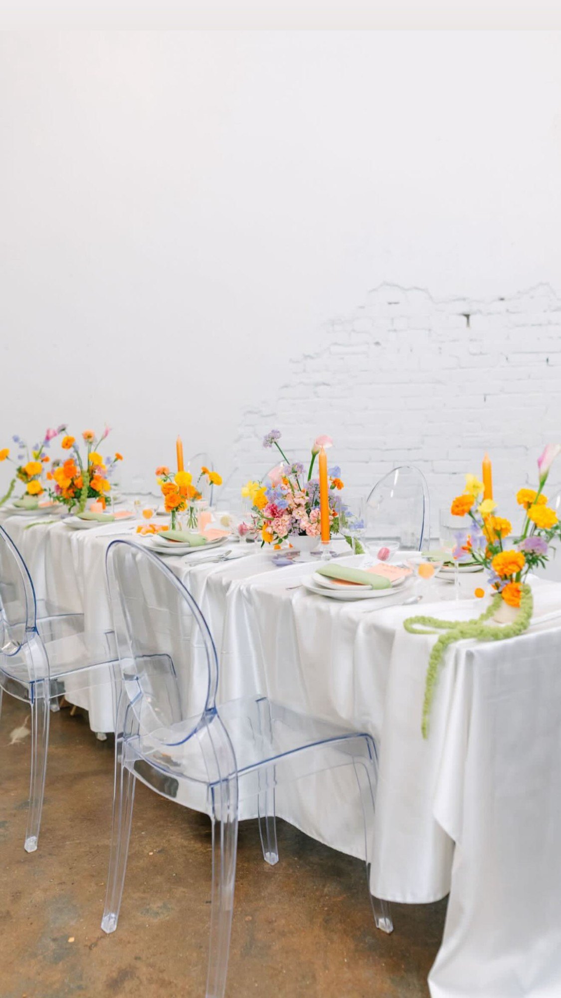 A long white satin table with ghost chairs, marigold arrangements and orange tapers