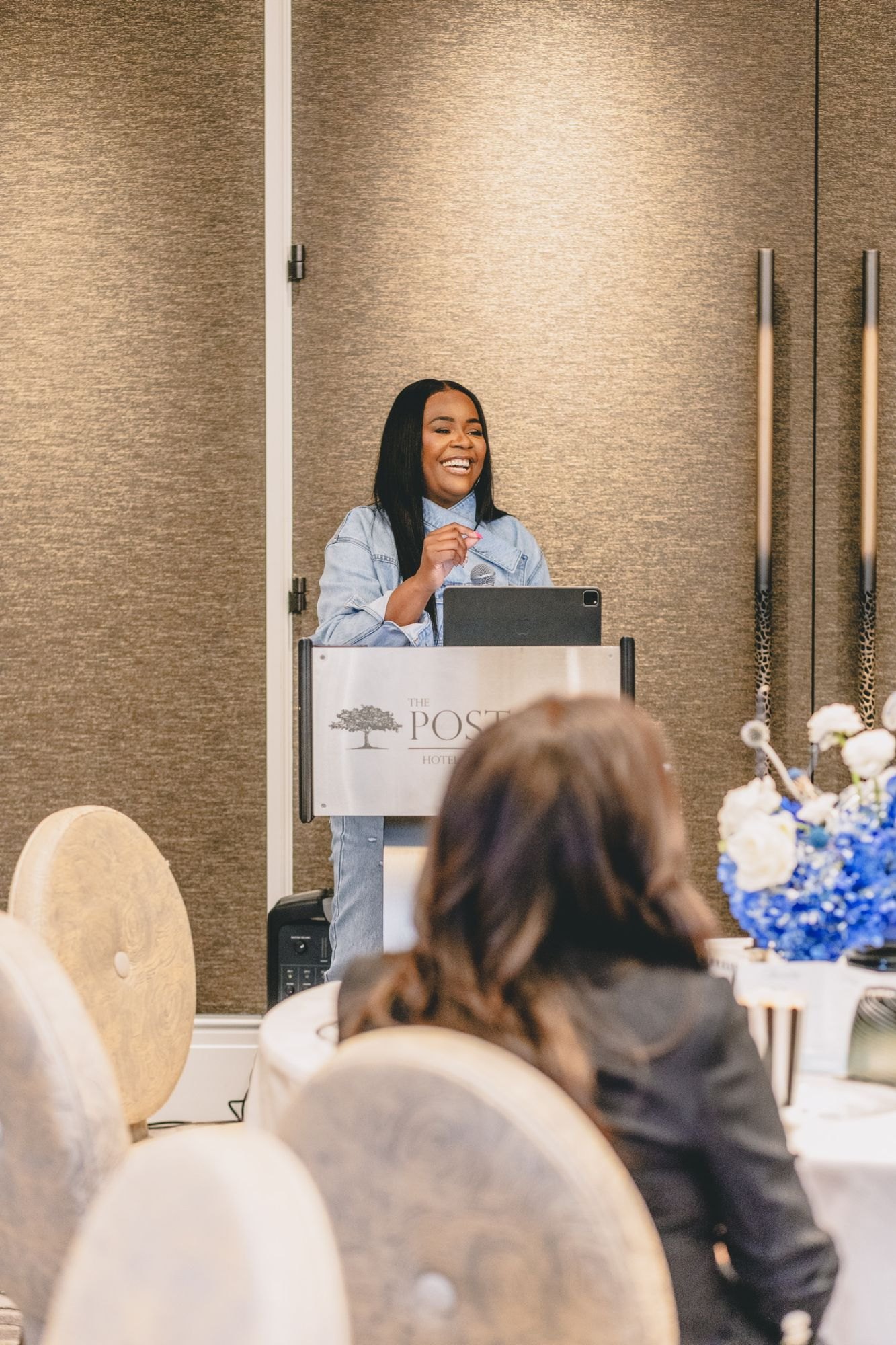 A speaker smiles from the podium in front of the blue and white tables